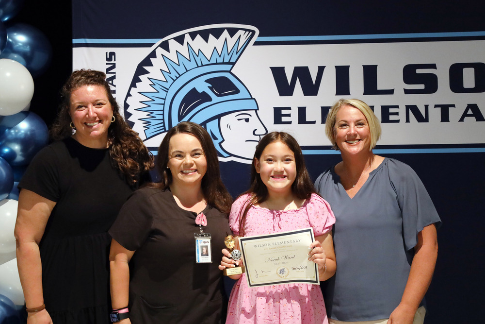 Three women and student hold certificate in front of Wilson Elementary School banner