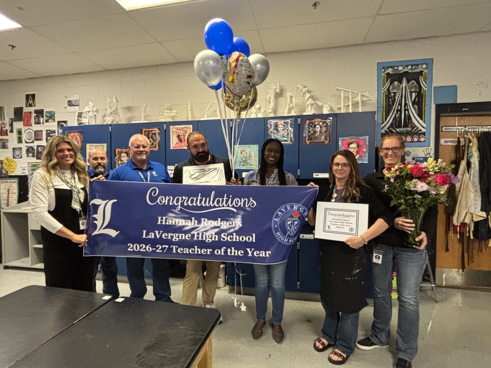 photo of Hannah Rodgers surrounded by the 6 administrators holding a banner congratulating her for being teacher of the year, coach jones is holding a cake, Dr. Hatchett has a blue and silver balloon bouquet, Dr. Hodges is holding a bouquet of seasonal flowers, and Mrs. Rodgers is holding her framed award certficate. Her classroom cupboards are in the background 