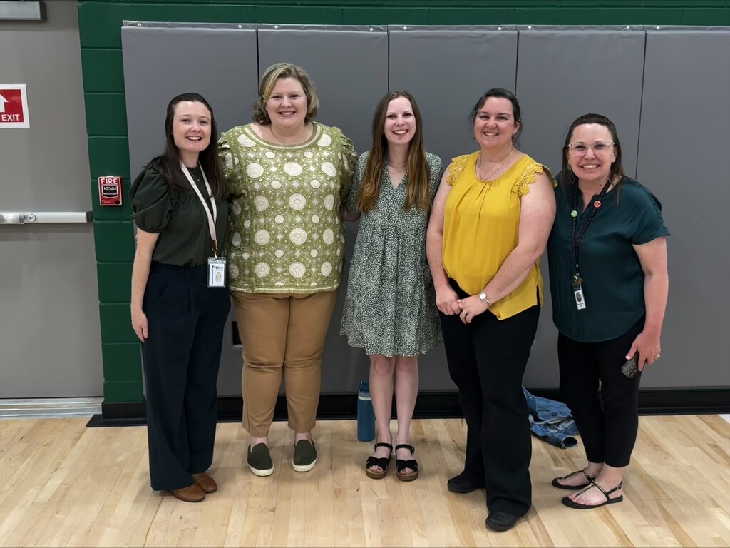 Five women in different colored shirts stand together in a room with a green wall and a wooden floor.