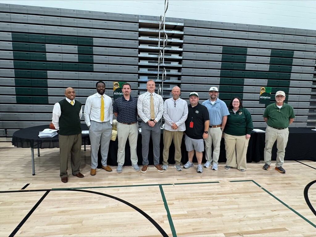 Nine people stand on a basketball court, smiling for a photo, with a large "H" on the wall.