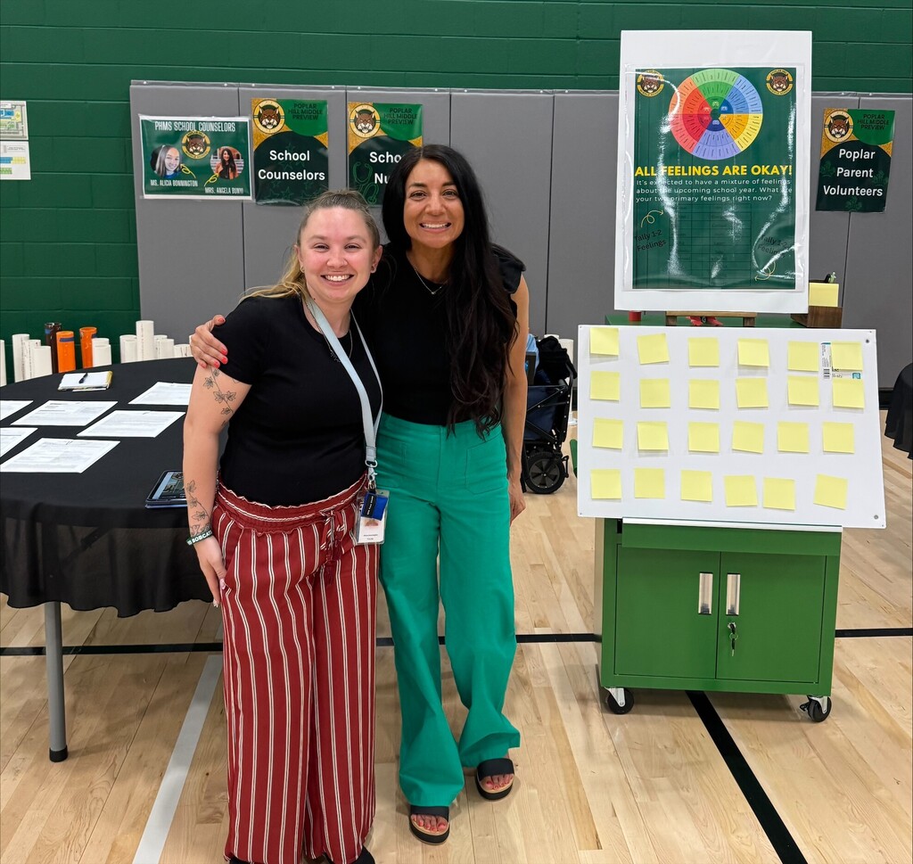 Two women stand near a colorful poster and table with sticky notes, in a room with wooden floors.