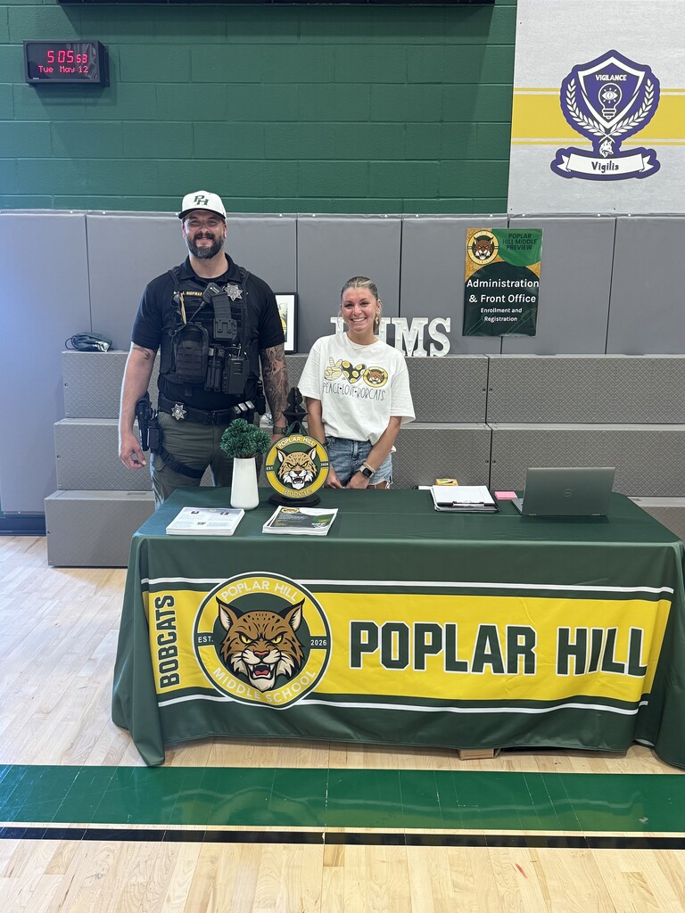Two individuals stand at a table in a gym with a green tablecloth. A police officer stands with a potted plant and a school logo.
