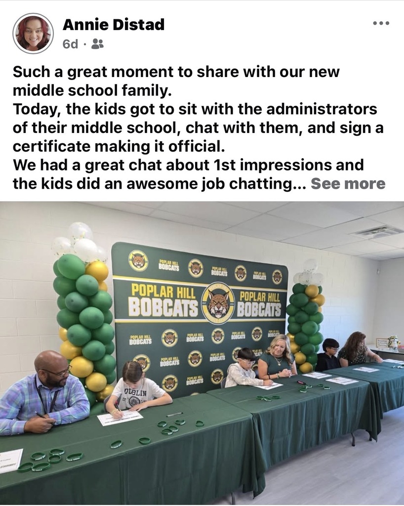 Several students and adults seated at a table with green backdrop, green balloons, and logos.