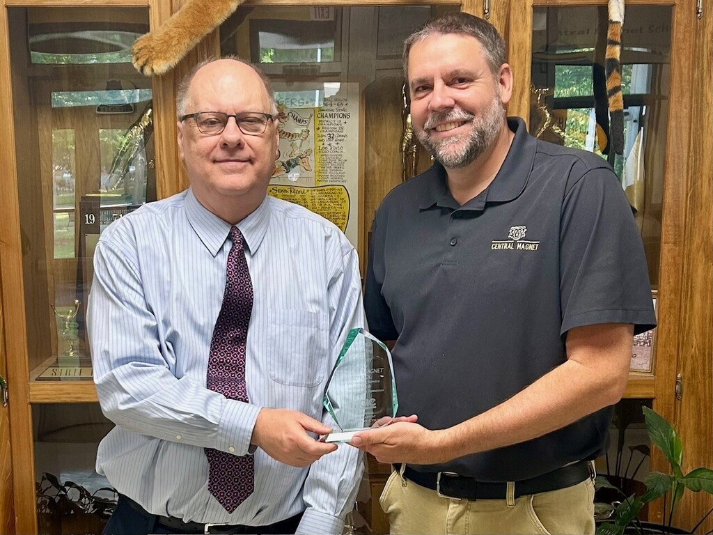 Two men stand indoors with a wooden display case behind them; one holds an award while the other smiles.