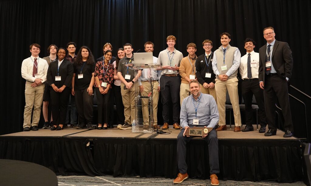 A group of young adults in formal wear standing on a stage, one seated with an award.