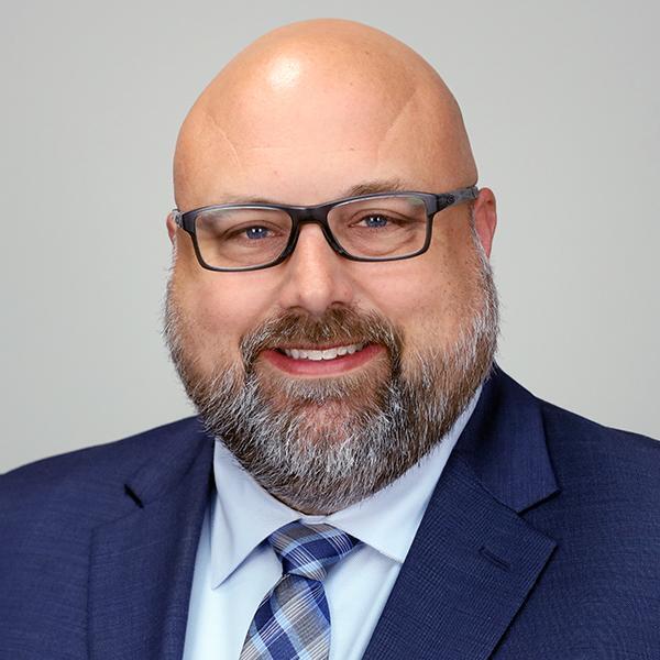 Man with a beard and glasses smiling, wearing a dark blue suit jacket, light blue shirt, and a checkered tie.
