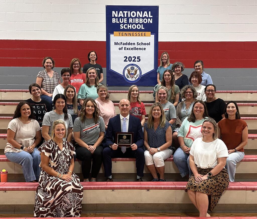 Group of women and one man sit on bleachers with a National Blue Ribbon School banner behind them.