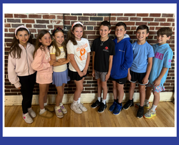 Seven children stand together in a group, against a brick wall. They wear casual clothing. Floor is wooden.