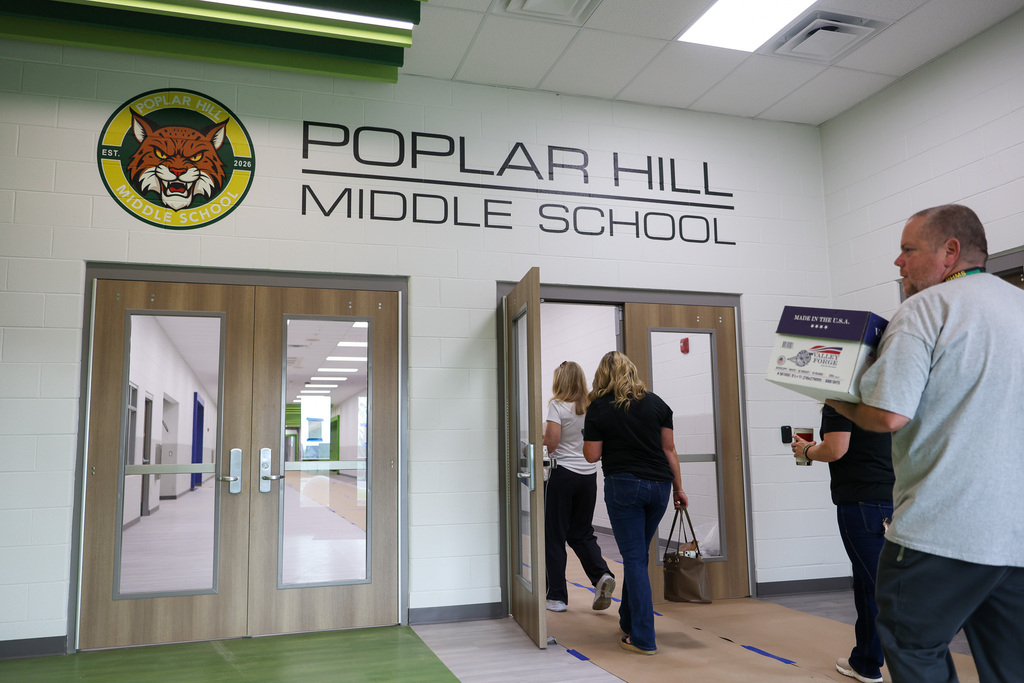 Three people enter a building with "Poplar Hill Middle School" on the wall. Two people carry boxes, one holds a handbag.