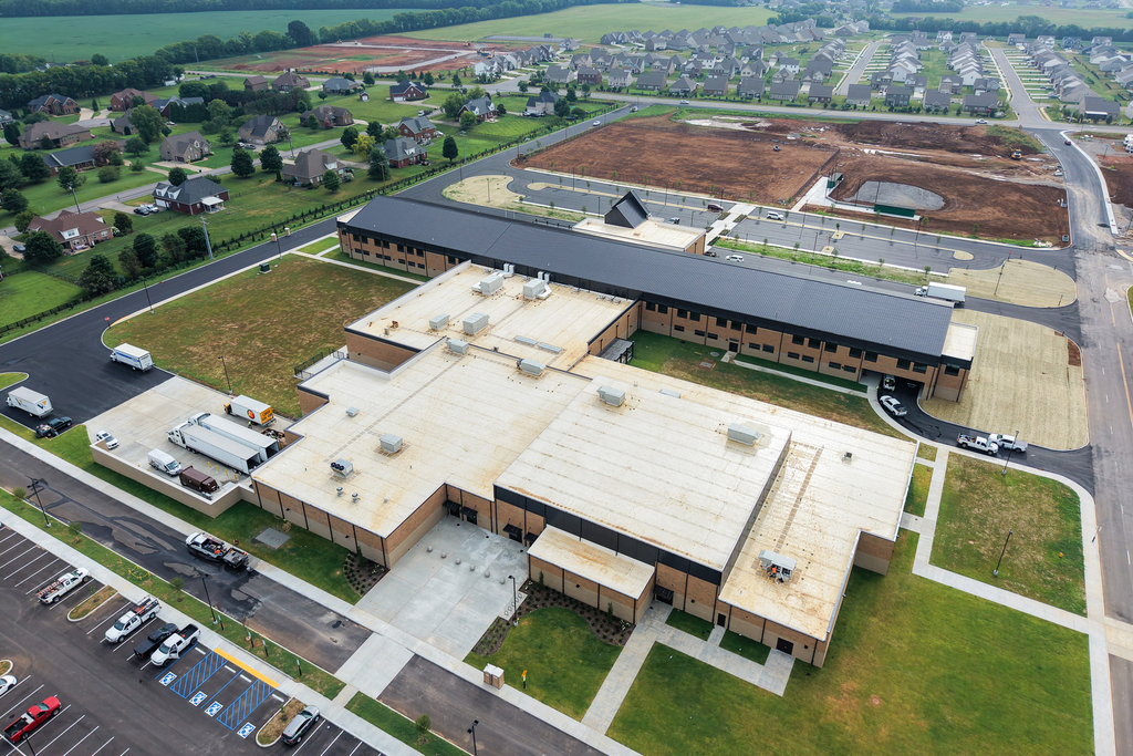 A large building with a parking lot. Many vehicles parked, some with trucks. Residential area in background.