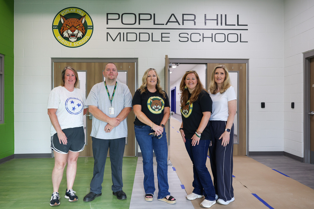 Five people stand in a row outside a school entrance. They wear casual clothing and sneakers.