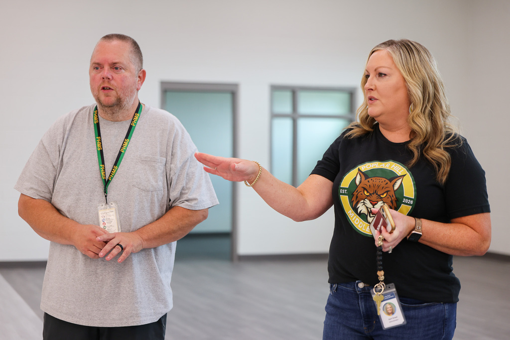 Two people stand in an indoor space with white walls and a gray floor; one wears a gray shirt and the other a black shirt.