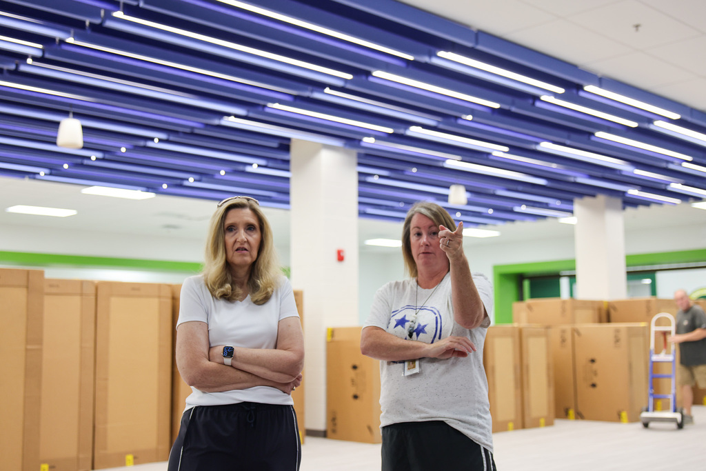 Two women in white shirts and black pants stand in a warehouse with blue lights. They face each other.