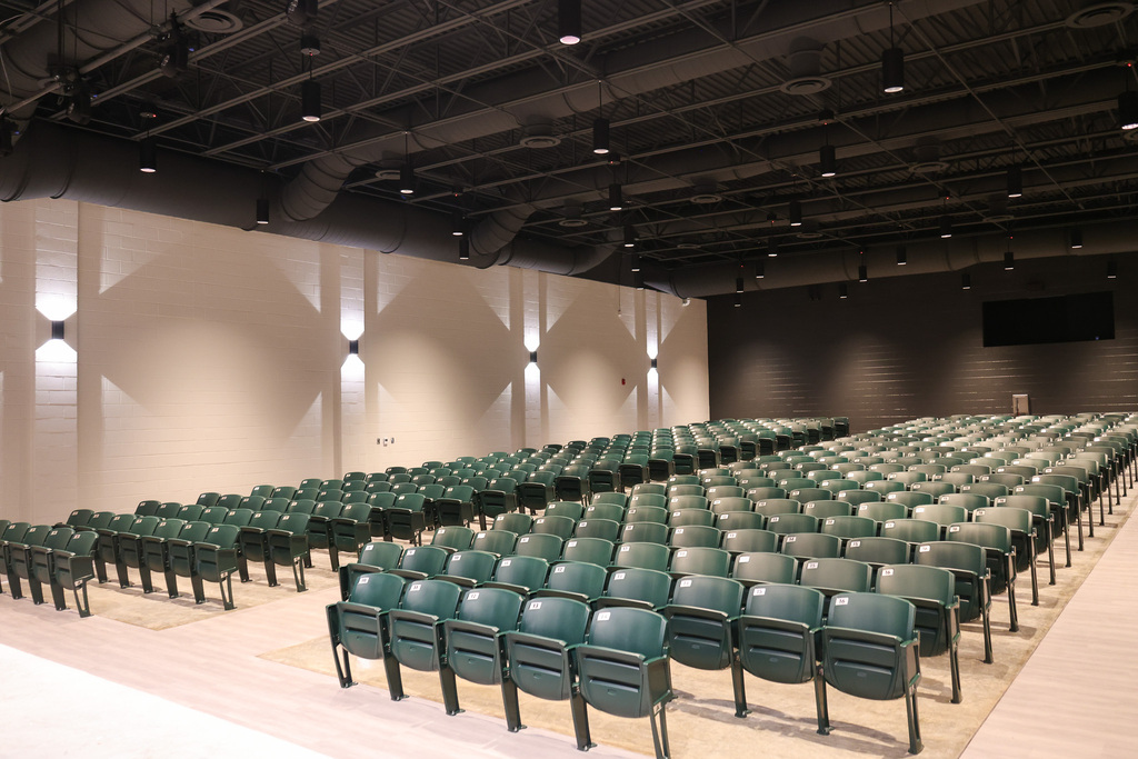 An auditorium with green chairs arranged in rows, white walls, and recessed lighting along the wall.