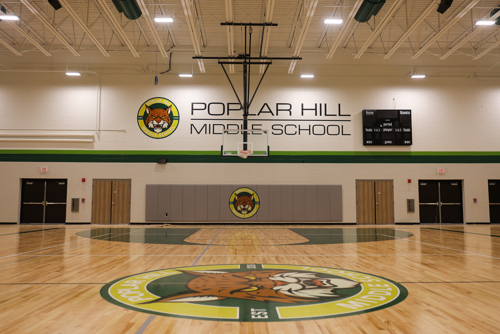 Indoor basketball court with a wooden floor. Walls feature school logo and name. Two doors and a scoreboard.