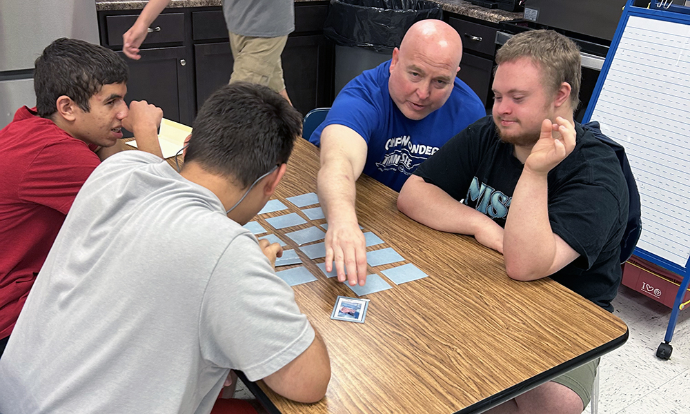 Four individuals seated around a table, engaged in a card game. Two males wear glasses, one in blue, one in gray.