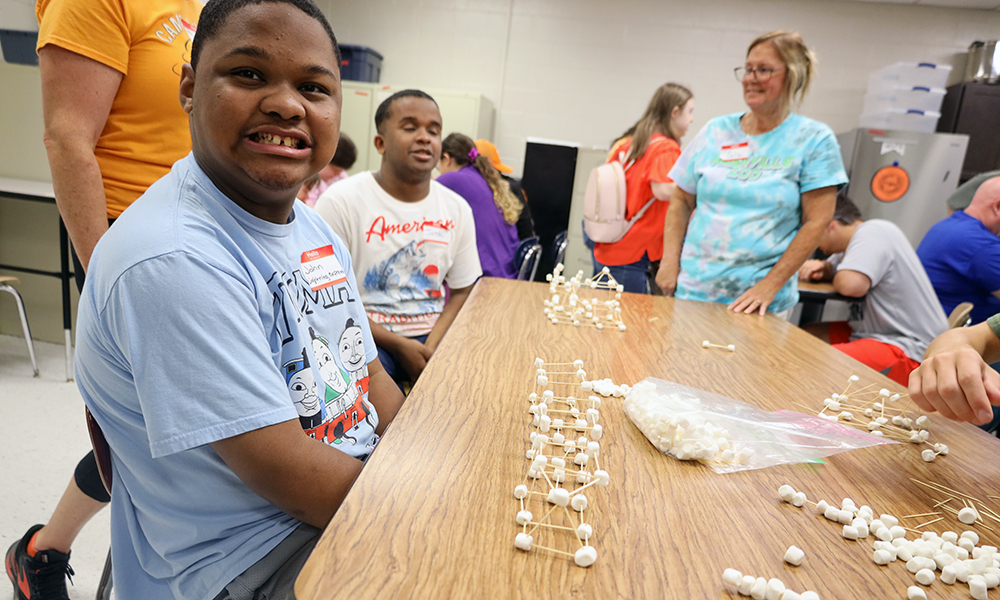 A person in a light blue shirt sits at a table with model building materials.