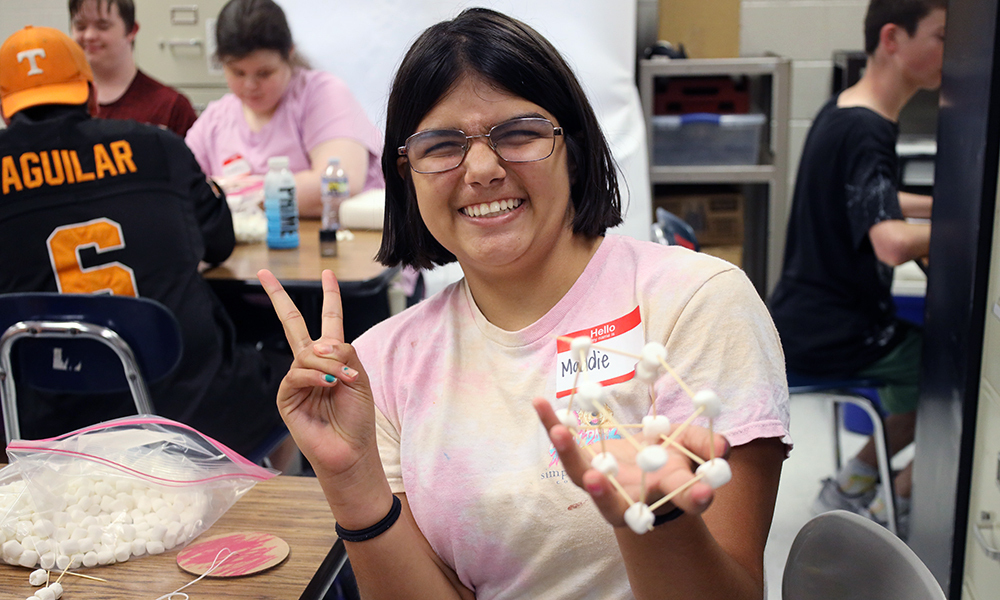 A person in a pink shirt holds a molecular model in a classroom with others working at tables.