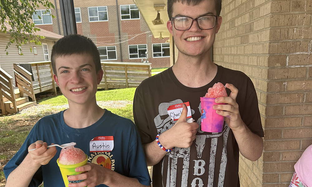 Two young boys hold slushy cups; one smiles with a thumbs up. Background features buildings and greenery.