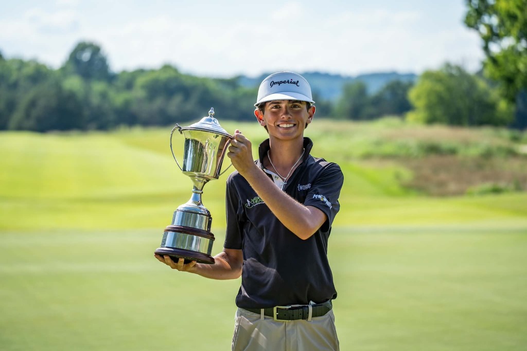 A young man poses with a trophy on a golf course, wearing a hat, a black shirt, and khaki pants.