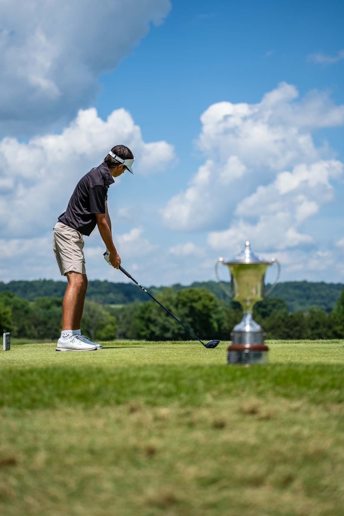 Golfer in shorts swings club on a green field; a trophy is behind him. Background has trees and a cloudy sky.