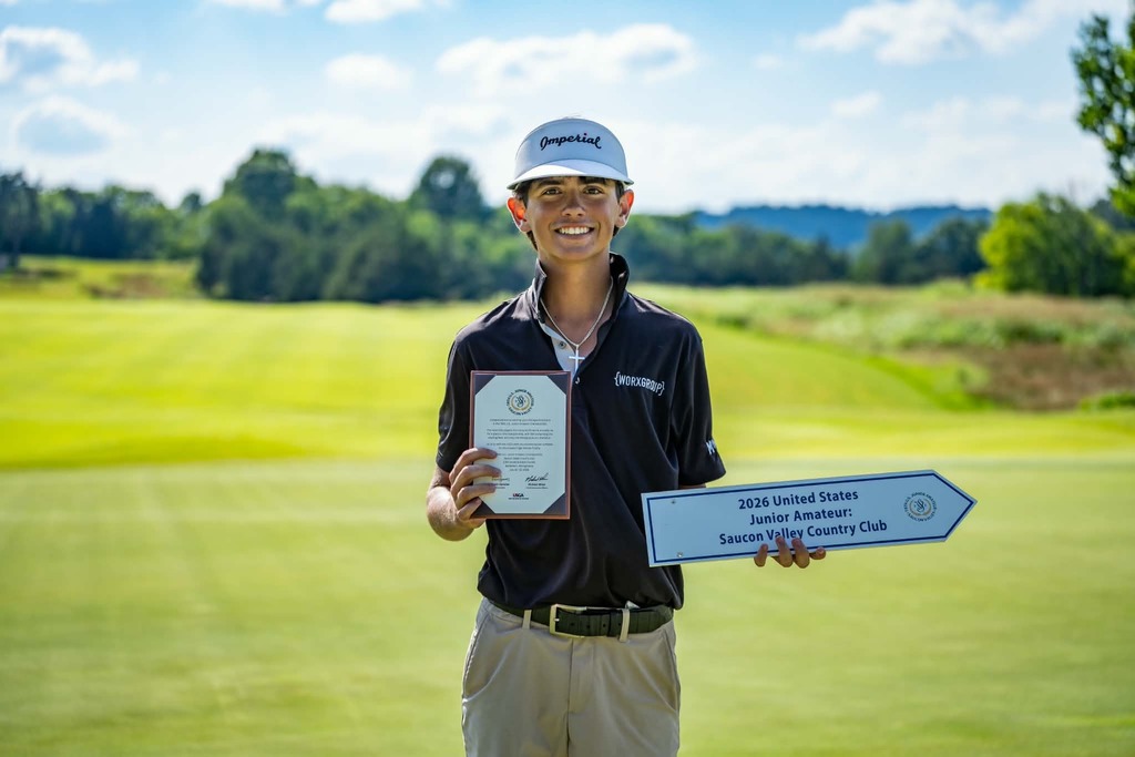 Person wearing a hat and holding a certificate and sign on a golf course.