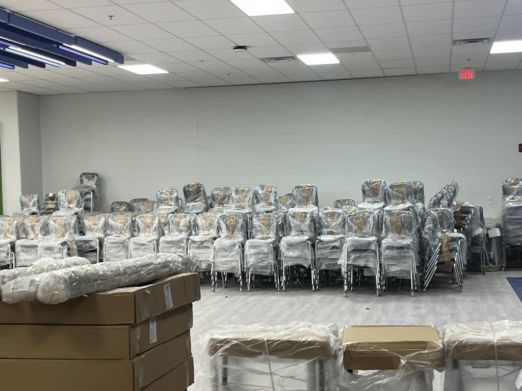 A room with rows of chairs covered in plastic. Boxes are in front and along the wall. White walls and ceiling with recessed lighting.