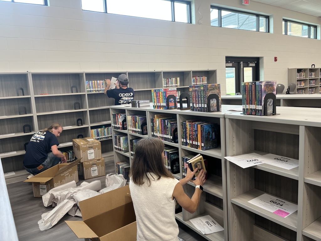 In a library, several people are arranging books on shelves, with a few boxes on the floor.