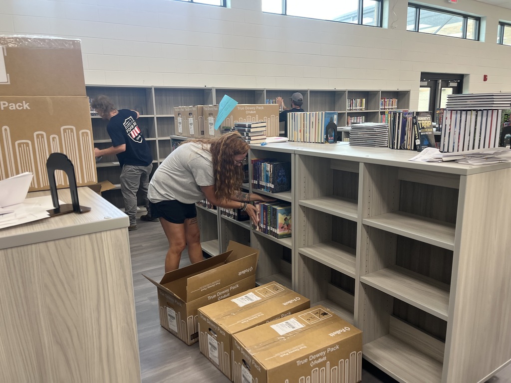 Two people in a library working on boxes and organizing books on shelves. Some boxes are on the floor.