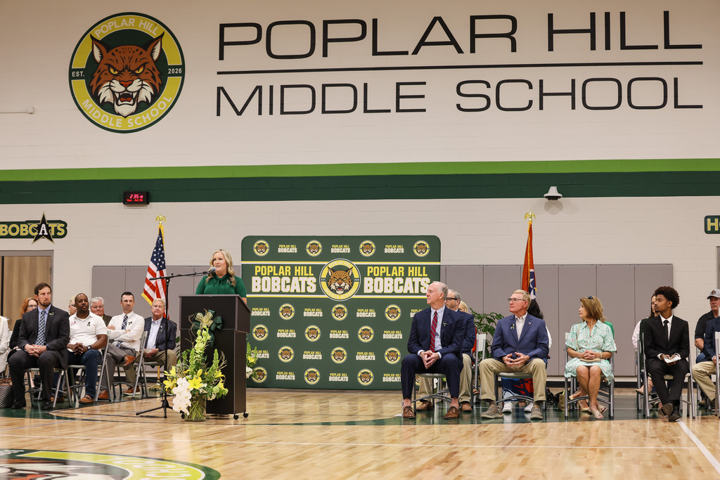 Speakers at a podium in a gymnasium with a banner that says "Poplar Hill Middle School" in the background.