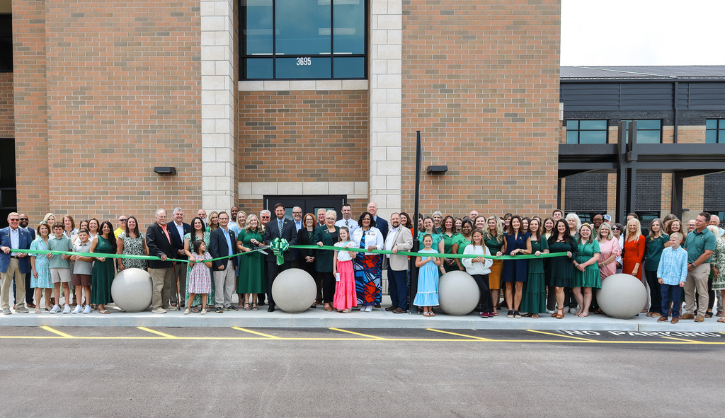 A group of people stands before a brick building, holding a green ribbon for a ribbon-cutting ceremony.