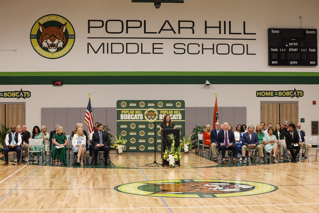A school gymnasium with a stage, microphone, and podium. People seated in chairs and standing behind the stage.