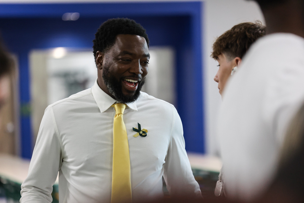 A man with a beard wearing a white shirt and yellow tie talks to another person in a blue-walled room.