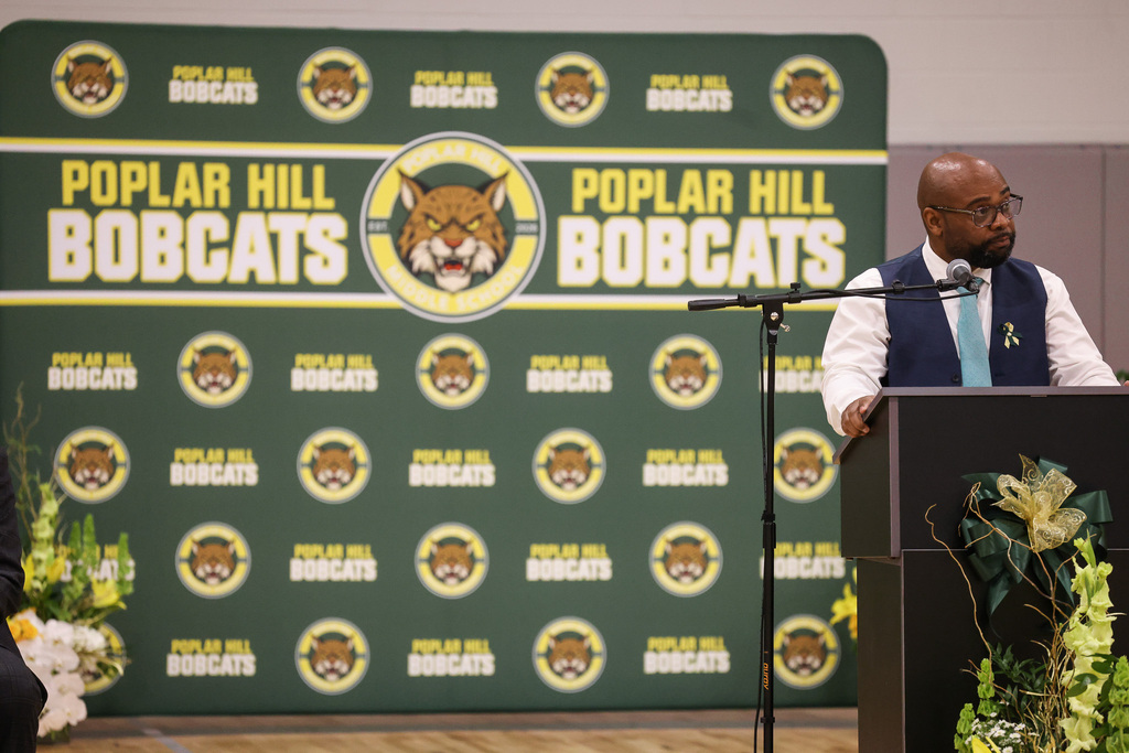 A podium with flowers stands in front of a person who speaks into a microphone. Behind them, a green backdrop displays "Poplar Hill Bobcats" with logos.