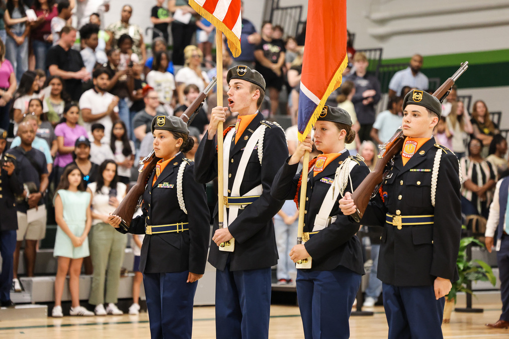 A group of young cadets in uniforms stand in a gym with flags. Audience seated in bleachers.