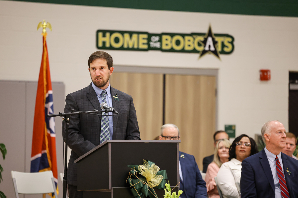 Man in suit at podium with microphone, next to a flag, and a crowd of people.