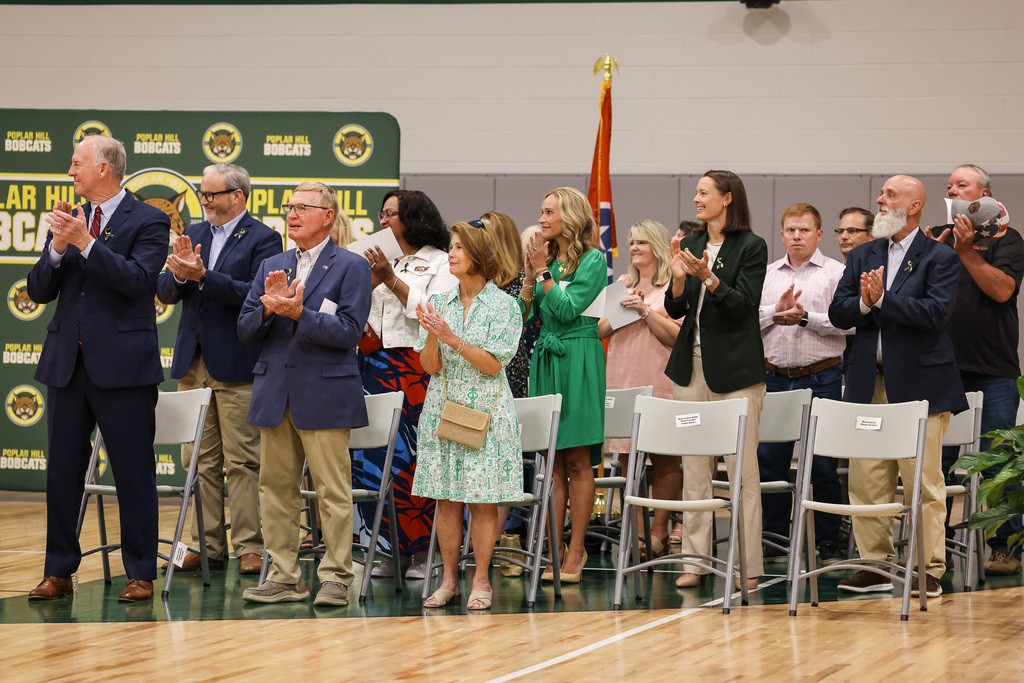 People in business attire stand on a basketball court in front of chairs, clapping hands. A flag is behind them.