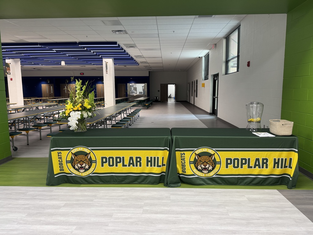 A school cafeteria with long tables and benches. Two tables are covered with green and yellow tablecloths. The tables have the words "Poplar Hill" written on them.