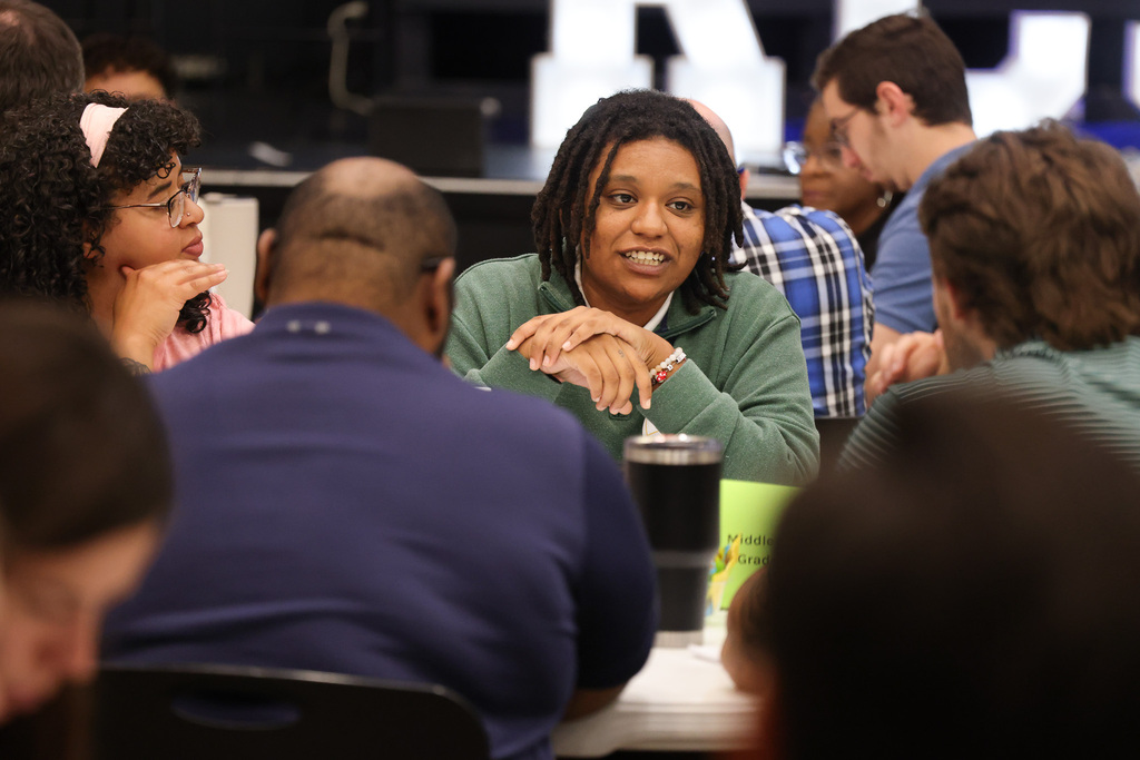 A group of people seated around a table; a woman wearing a green sweater speaks while others listen.