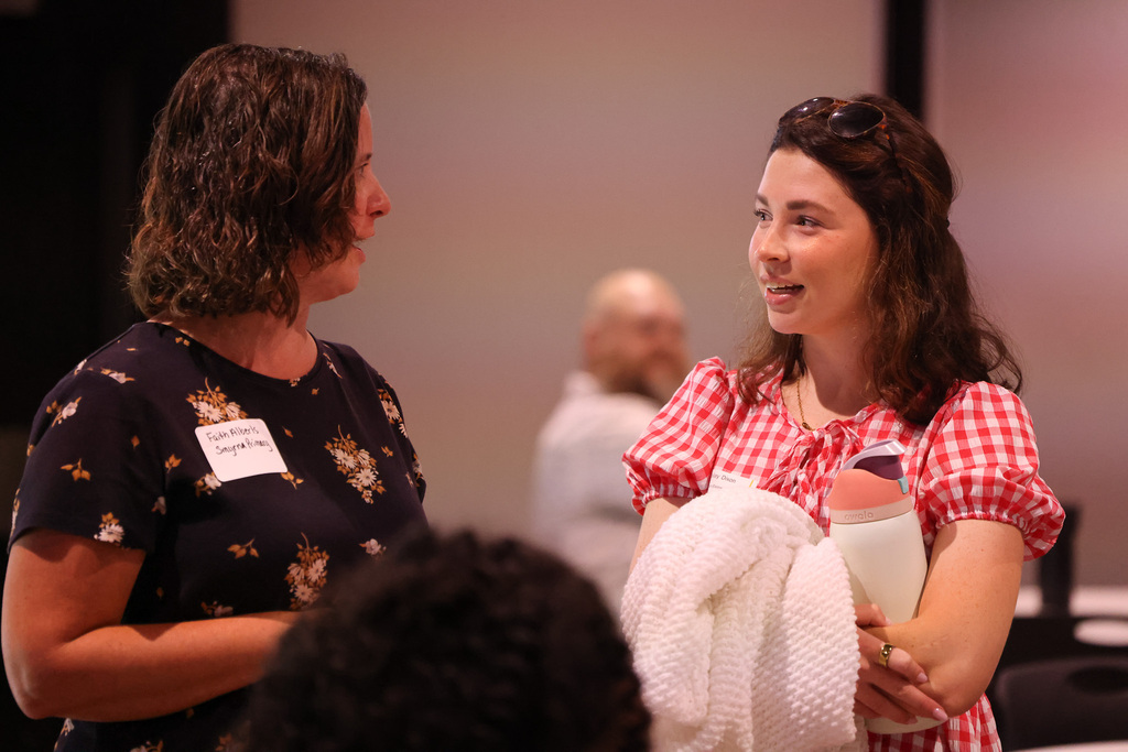 Two women converse indoors. One holds a baby and a towel. A third person stands in the background.