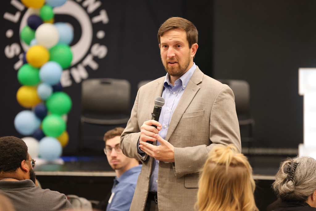 A man with a microphone speaks at an event. Behind him, a backdrop features a balloon arrangement and text.
