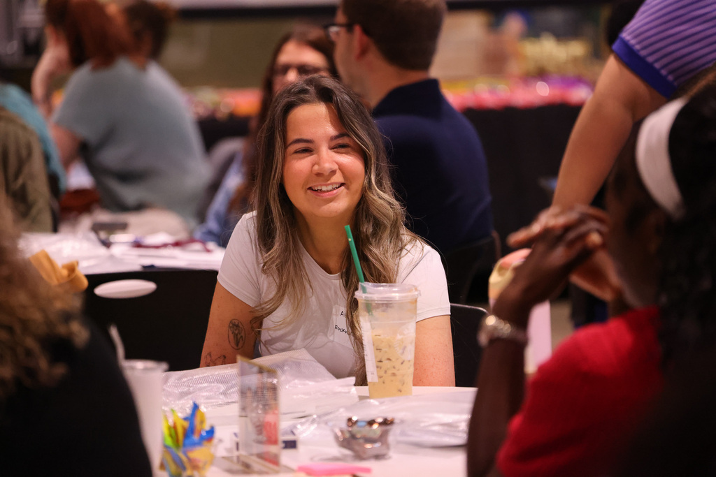A smiling woman with a straw in a drink sits in a chair at a table.