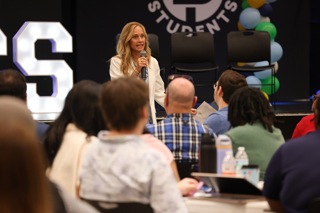 A woman speaks into a microphone on stage. People sit in chairs around tables, listening. The word "CS" is on a lit sign.