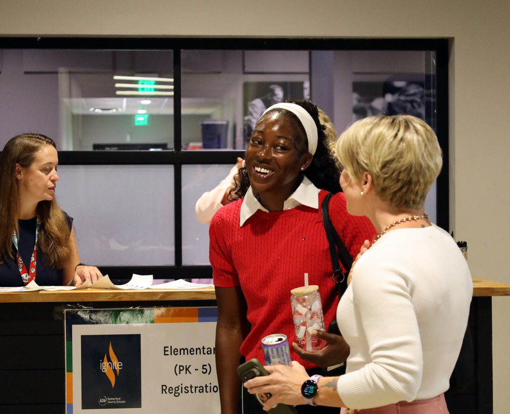 Three women stand in front of a counter; one holds a drink cup, another smiles, and the third talks to them.
