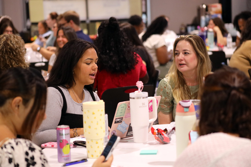 Two women at a table talk as one uses a computer.