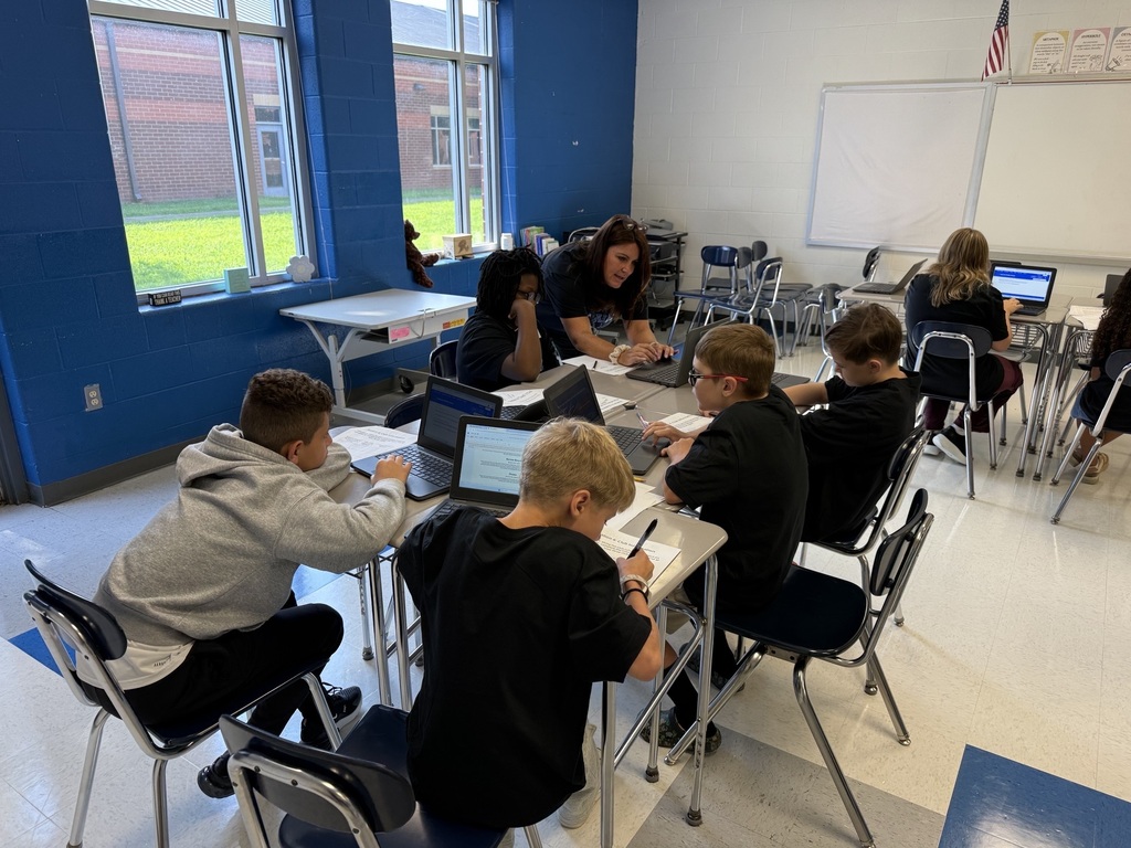 A classroom with blue walls, a whiteboard, and large windows. Students sit at desks, working on laptops.