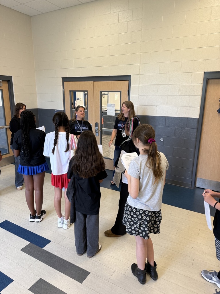 Students in a hallway converse with a teacher. Three doors, one open, are in the background.