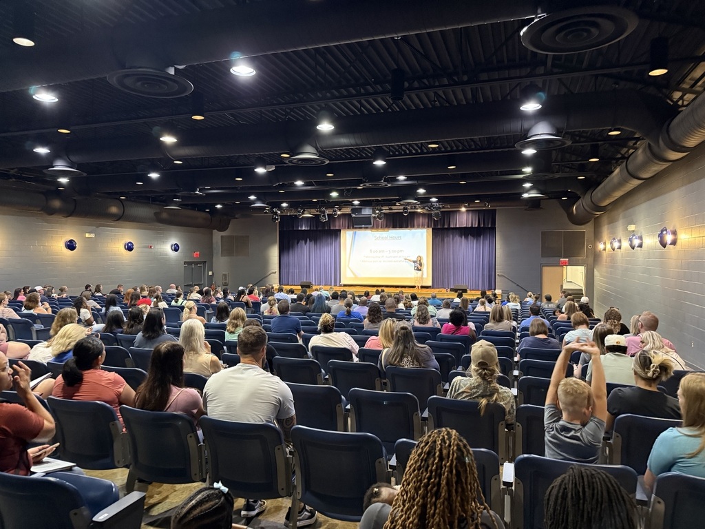 Large room with many people seated in rows facing a large screen, with lights on the ceiling.