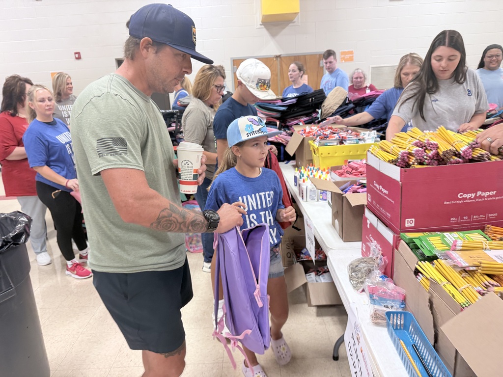 A man with tattoos helps a child with a purple backpack, surrounded by people in a room with tables full of supplies.