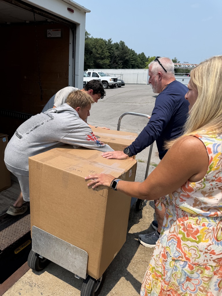 Four people handle boxes on a dolly in an outdoor area. A truck is parked in the background.